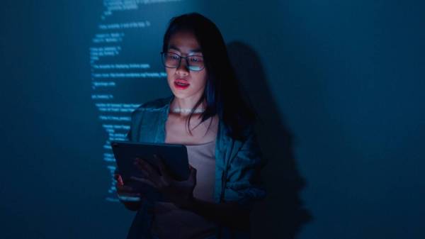 In this photograph, a young Asian woman is seen looking at a screen with lines of code projected over her face as she talks to a group of computer science students who are out of shot.