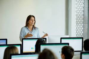 Photograph of female teacher teaching university students. Men and women are listening to professor in computer lab. They are sitting in classroom.