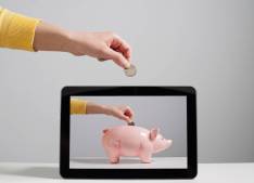 A photograph representing a 'digital piggy bank' showing a photograph of a hand about to drop a coin into a piggy bank displayed on a tablet screen, with the same image of a hand about to drop a coin duplicated above the tablet.