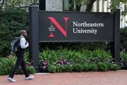 A Black male student wearing a pale blue shirt and backpack walks past a large wooden sign painted with the words "Northeastern University".